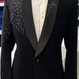 Black tuxedo with satin lapels and intricate embellishments on the left shoulder and chest, paired with a white shirt and black bow tie, displayed on a mannequin in a formal wear store.