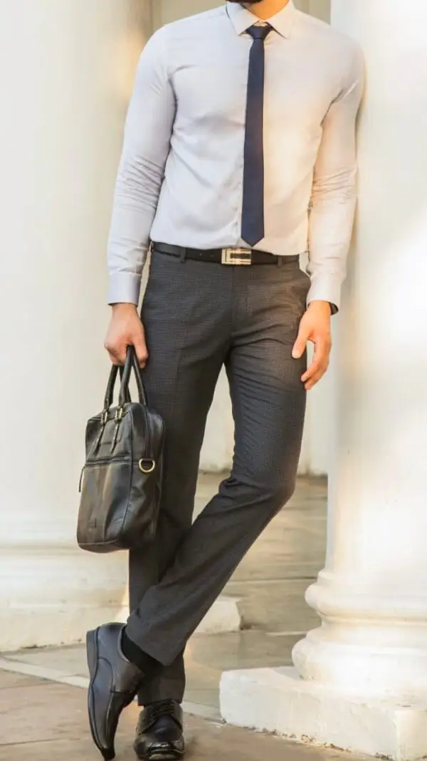 Man wearing a slim-fit white formal shirt, dark grey tailored trousers, and a navy blue tie, holding a black leather briefcase, exuding a professional and polished look suitable for office wear or formal occasions.